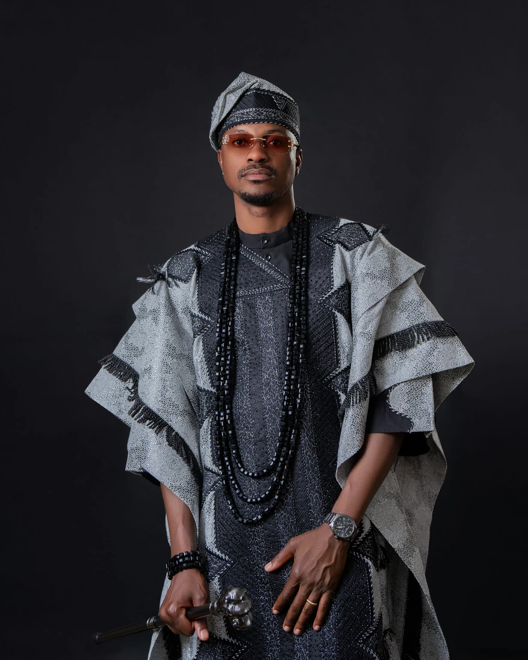 Close portrait of Philip Akinjobi wearing black-and-silver Yoruba attire and coral-tinted glasses